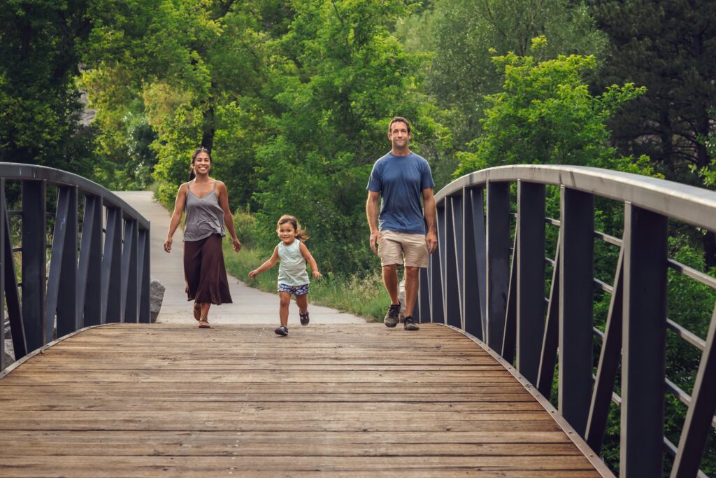 Father, mother, and their toddler daughter crossing over a bridge during their easy hike in Boulder, Colorado.