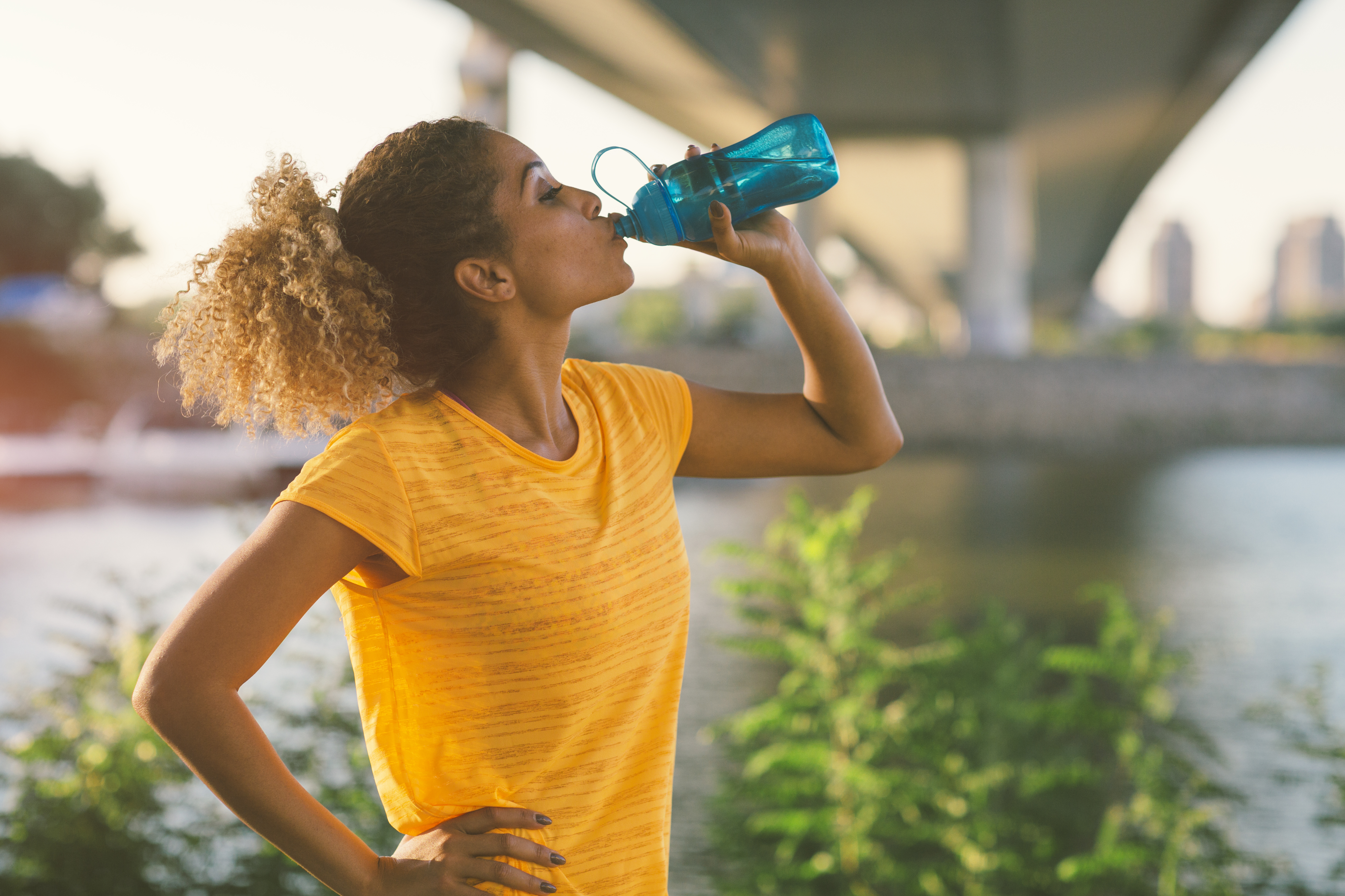 Latina Runner running in the city near river. Standing under the bridge, making break to drink some water. Cityscape in background.