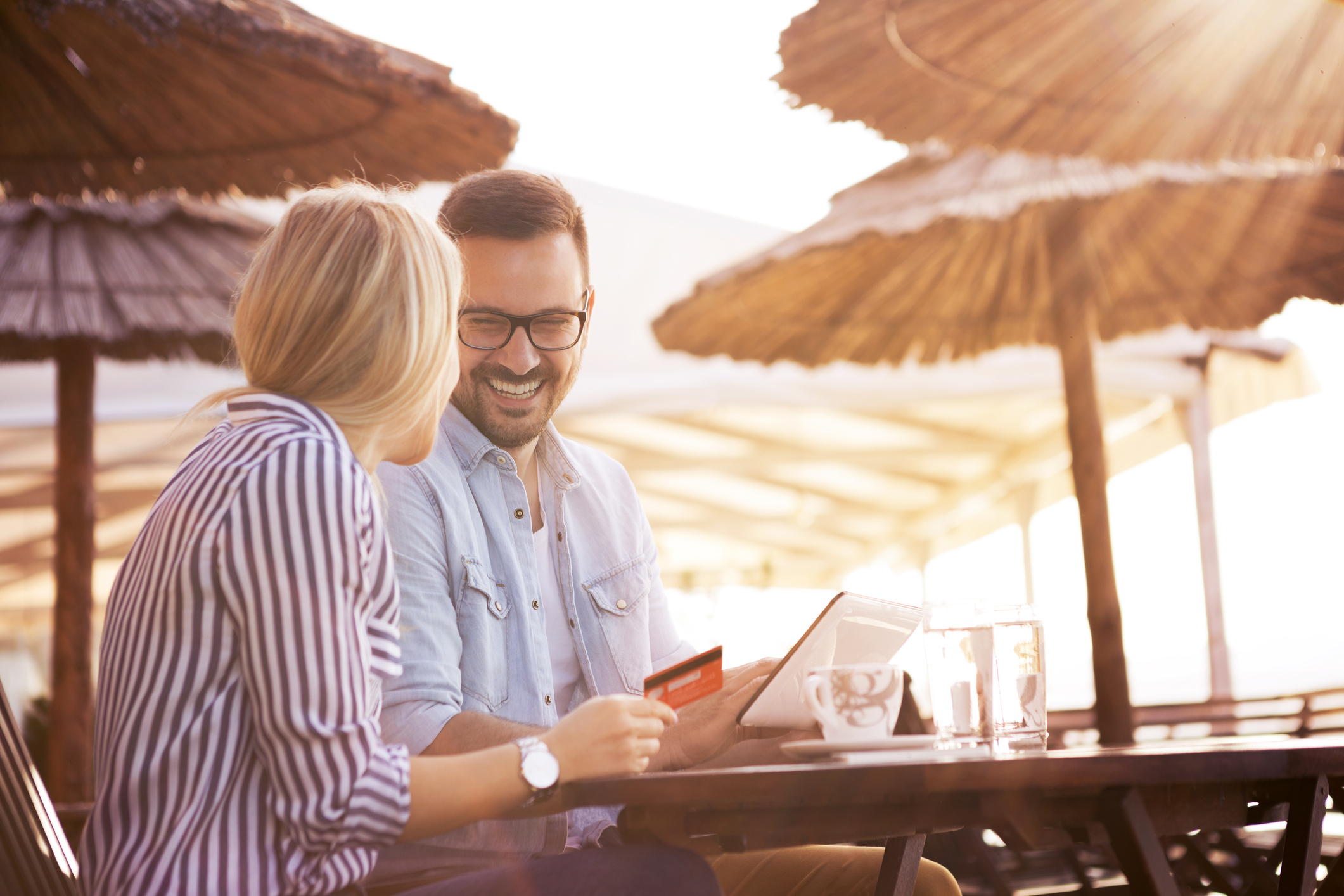 A handsome bearded young man and his pretty blonde girl sit in a restaurant on the seashore and they are happy because they bought something nice on the online shop using travel money card.