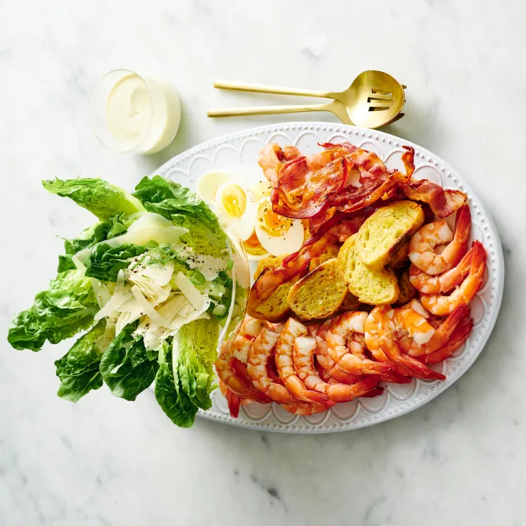 Plate of shrimp, bacon, lettuce, boiled eggs, croutons, and grated cheese with a fork, spoon, and dressing on the side.