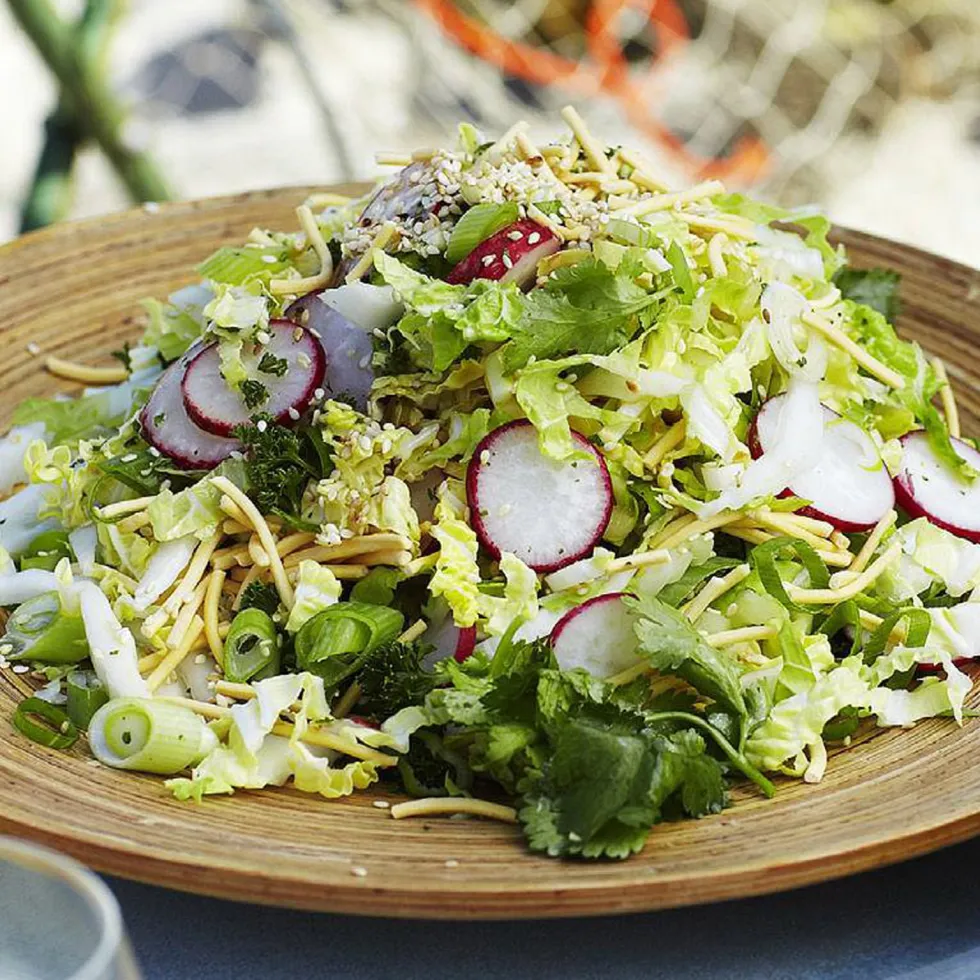 A wooden bowl filled with a fresh salad featuring sliced radishes, shredded lettuce, herbs, and sesame seeds.