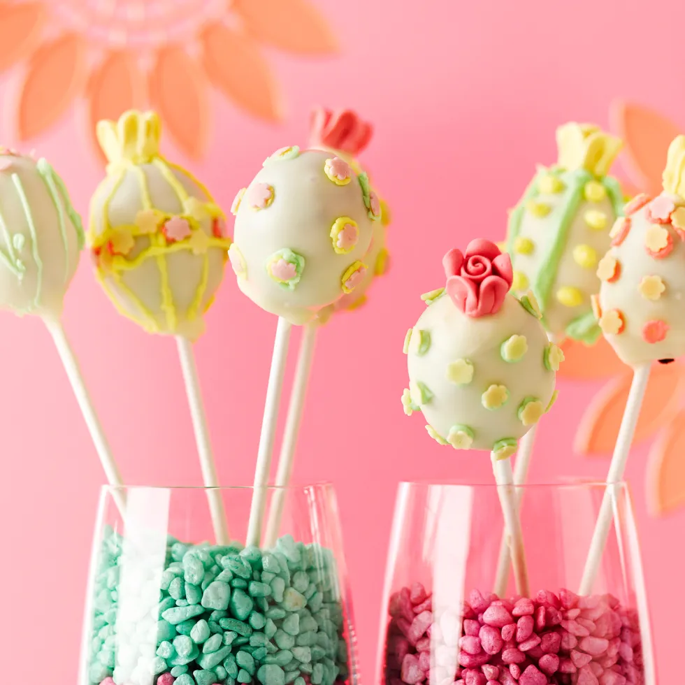 Colorful Easter egg cake pops with floral decoration displayed in glasses filled with candy rocks against a pink background.