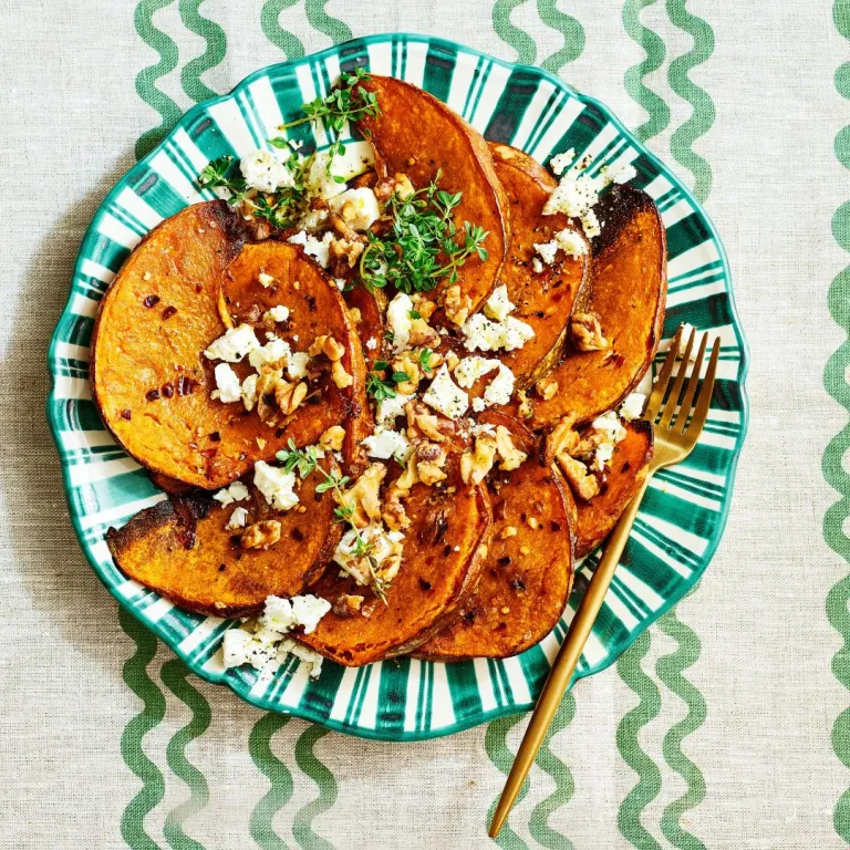 Roasted sweet potatoes topped with walnuts, feta, and herbs on a green striped plate, with a gold fork on the side.