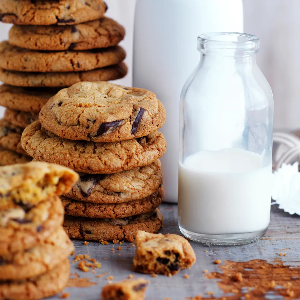 Stacks of chocolate chip cookies next to a bottle of milk with crumbs scattered on a wooden surface.
