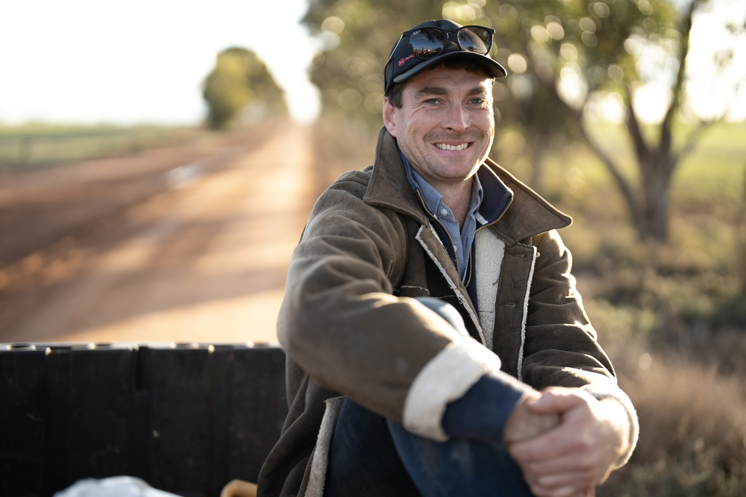 Man in warm jacket and cap sitting outdoors, smiling on a rural road with blurred trees in the background.