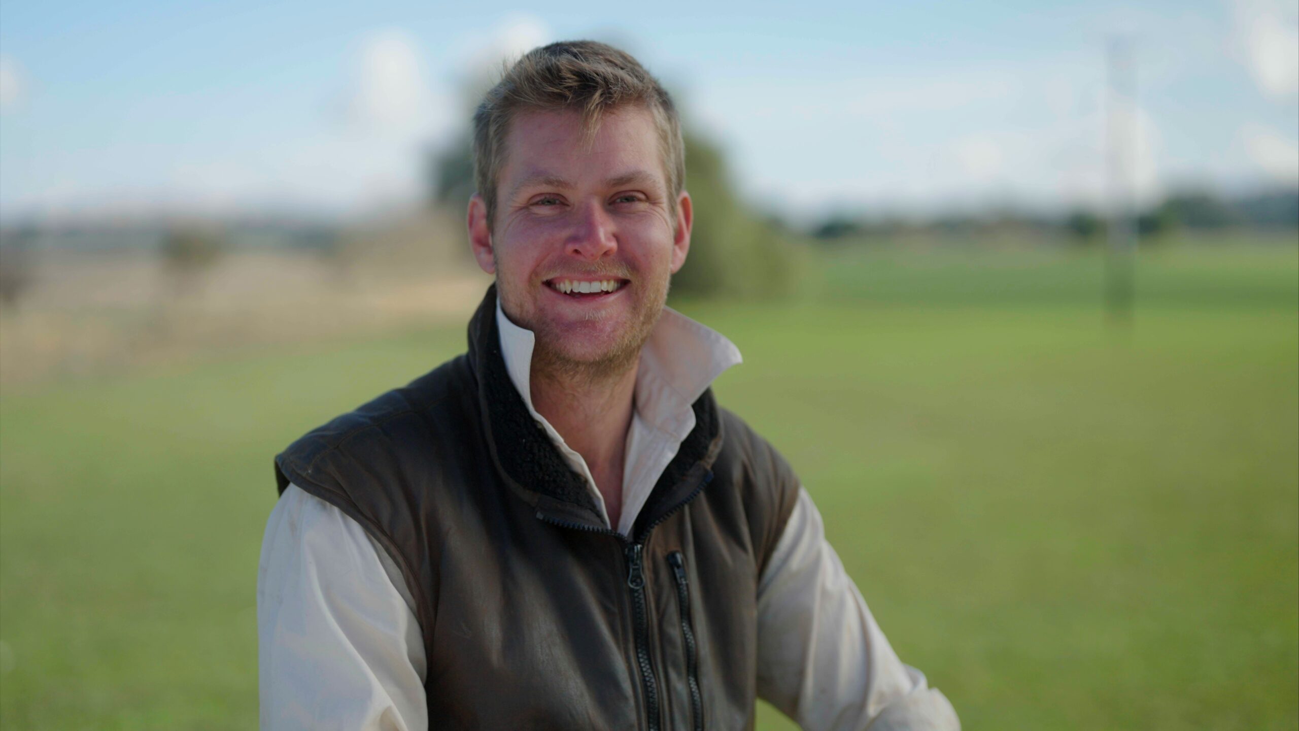 A man smiling outdoors with a blurred green field in the background.