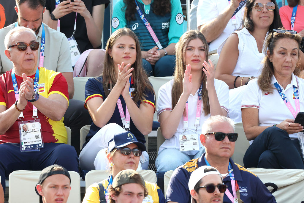 A group of people sitting in stadium seats, clapping and watching an event, wearing ID lanyards and sunglasses.