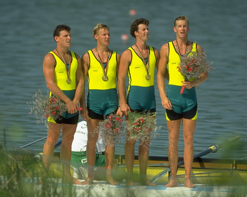 Four male rowers in Australia uniforms hold medals and flowers at a podium with water in the background.