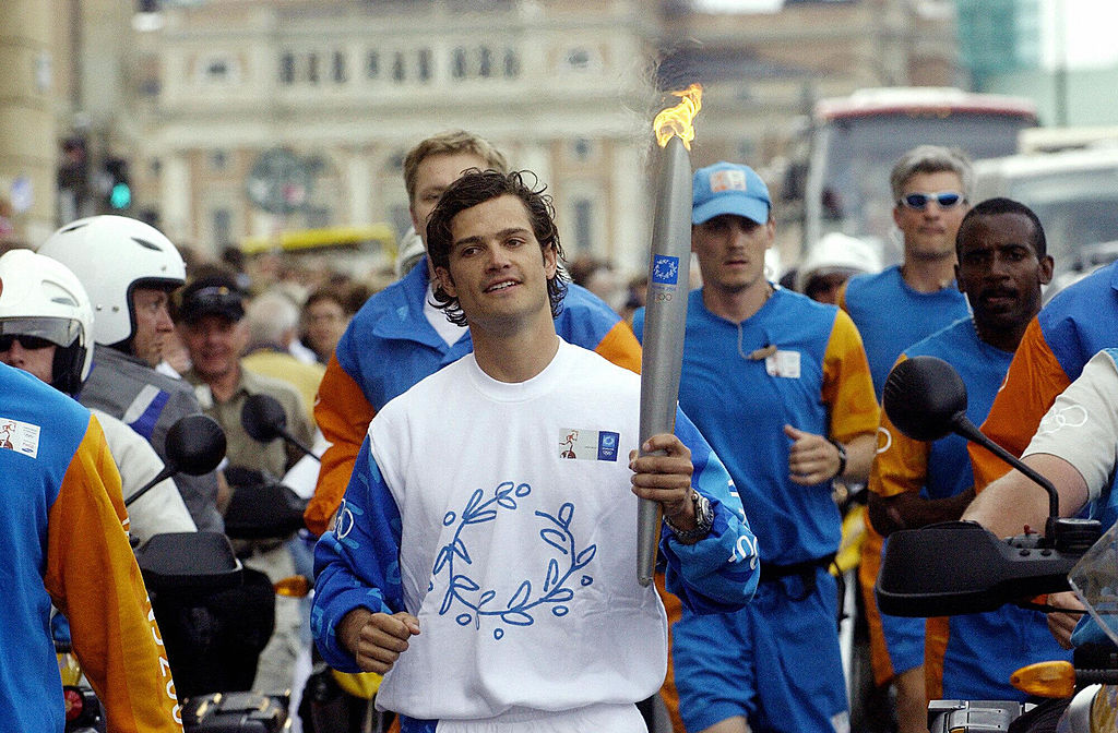 Olympic torchbearer running in city street, surrounded by security and onlookers, holding a lit Olympic torch.