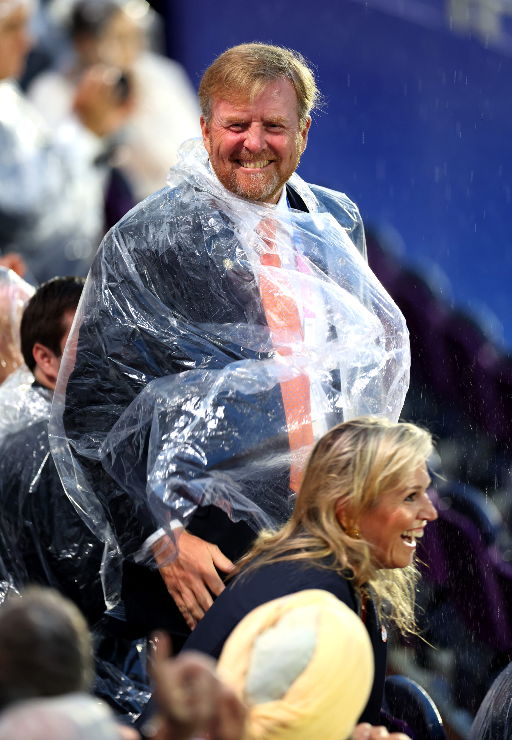 Man smiling in a rain poncho at an outdoor event, with people and colorful seats in the background.