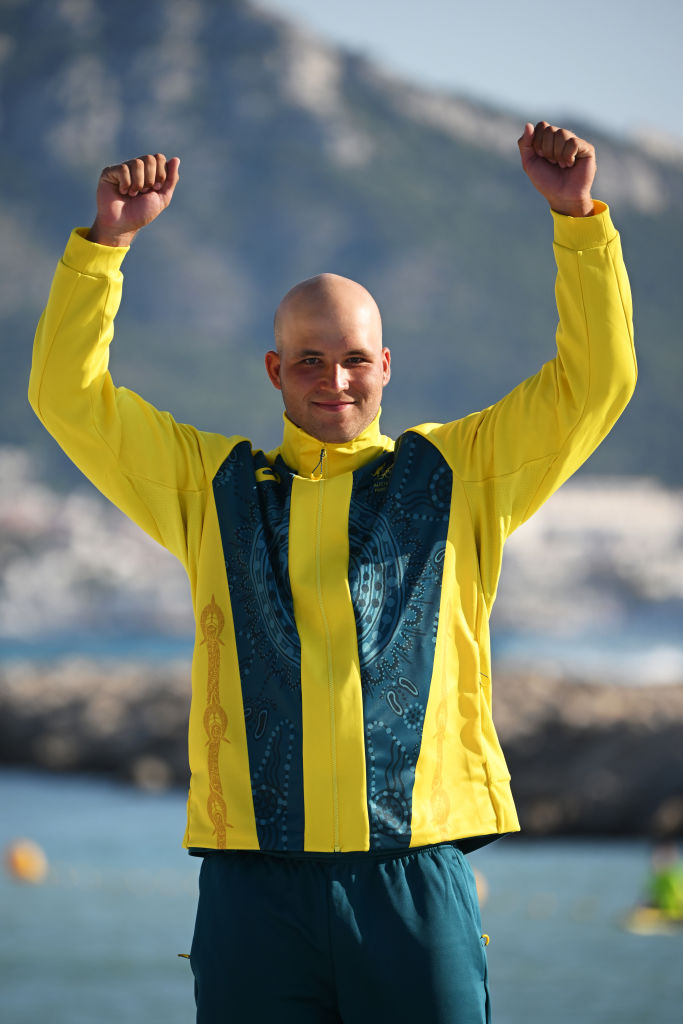 Athlete in a yellow and green jacket celebrates with raised arms, scenic mountain background.