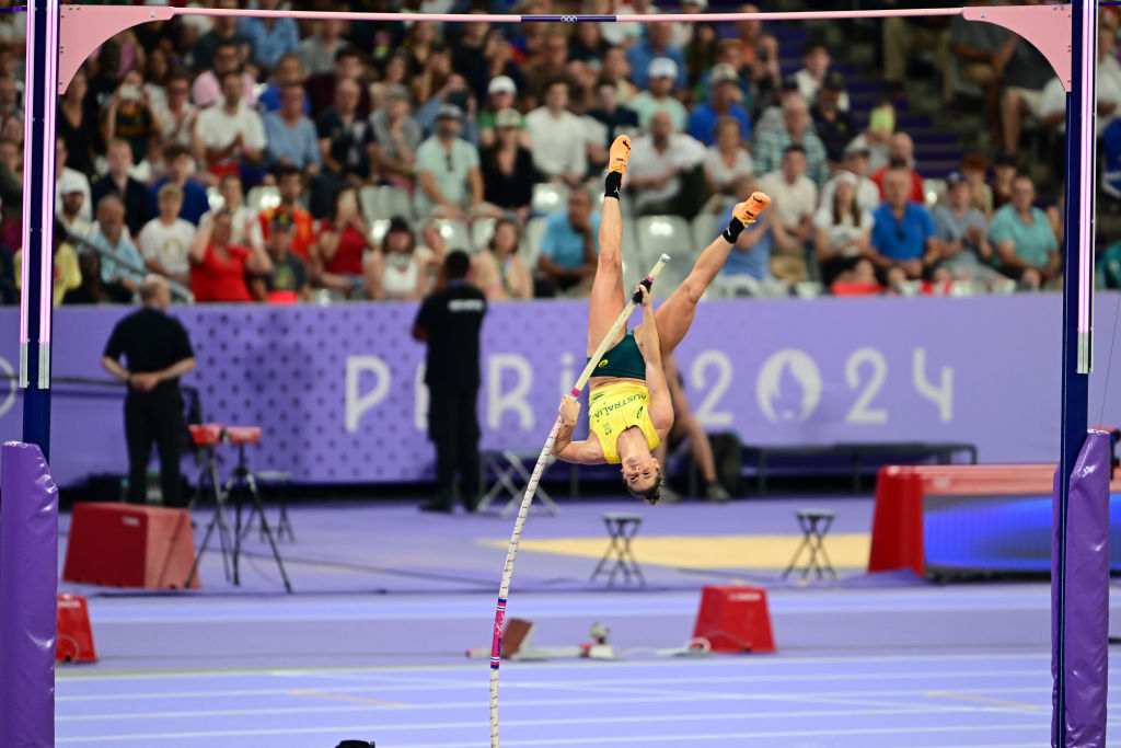 An athlete in mid-air during a pole vault event at a stadium with a crowd. Event is Paris 2024.