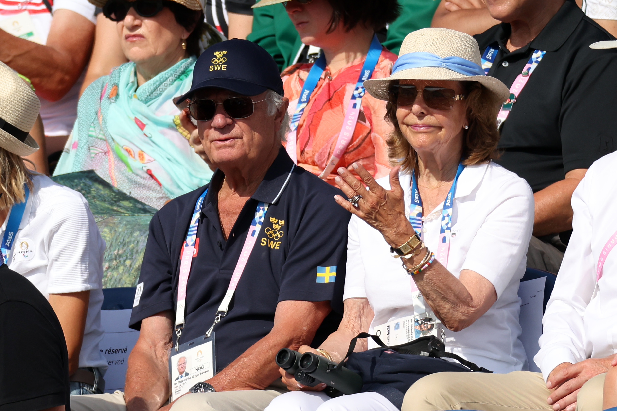 Two elderly individuals in sports attire and hats sitting in a crowd. One has a pair of binoculars.
