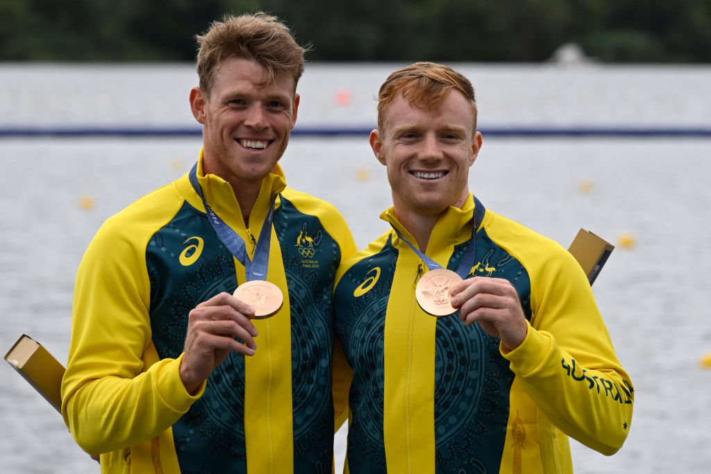 Two athletes in Australian tracksuits hold bronze medals, smiling with oars by a rowing venue background.