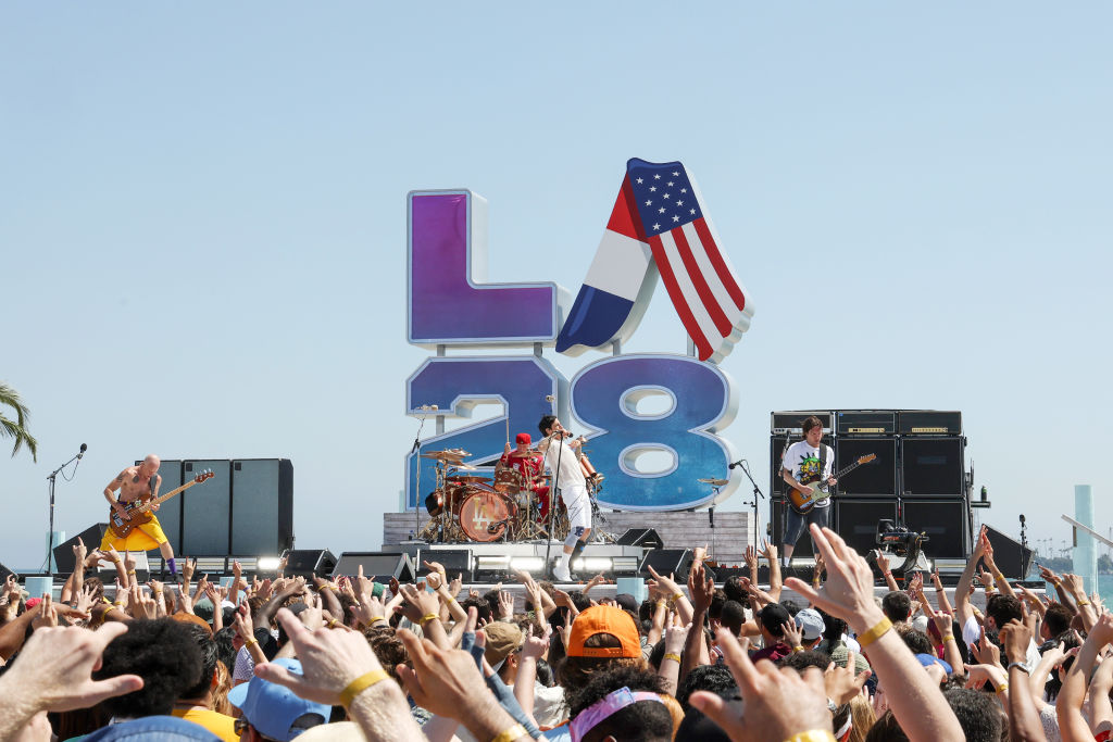 Band performing on stage with crowd in front, large "LA28" Olympic sign and USA flag, sunny outdoor event.