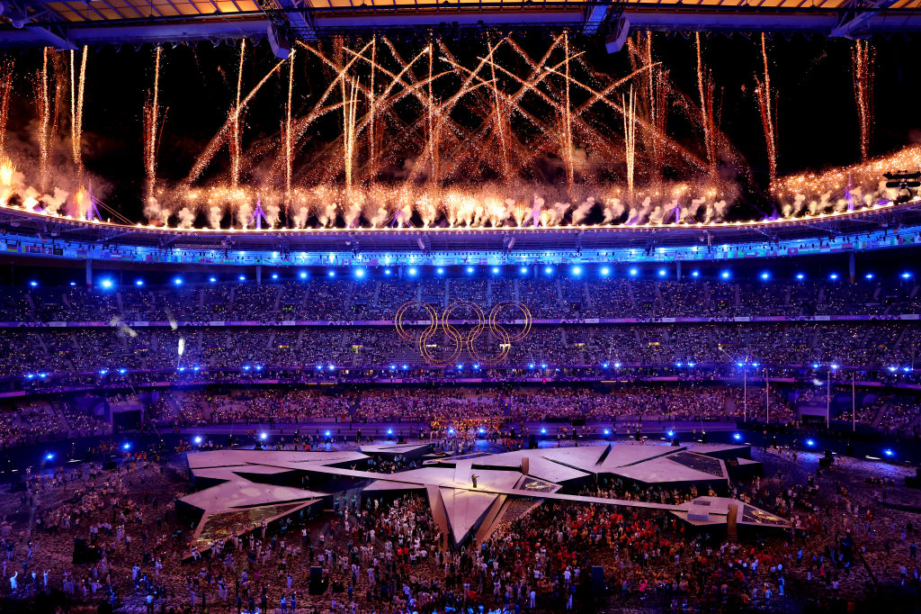 Olympic stadium with vibrant fireworks and glowing rings during the opening ceremony, crowded with spectators and performers.