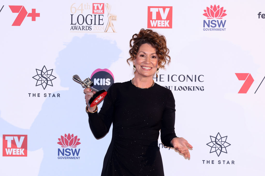 A woman in a black dress holding a Logie award, standing on a red carpet with sponsor logos in the background.