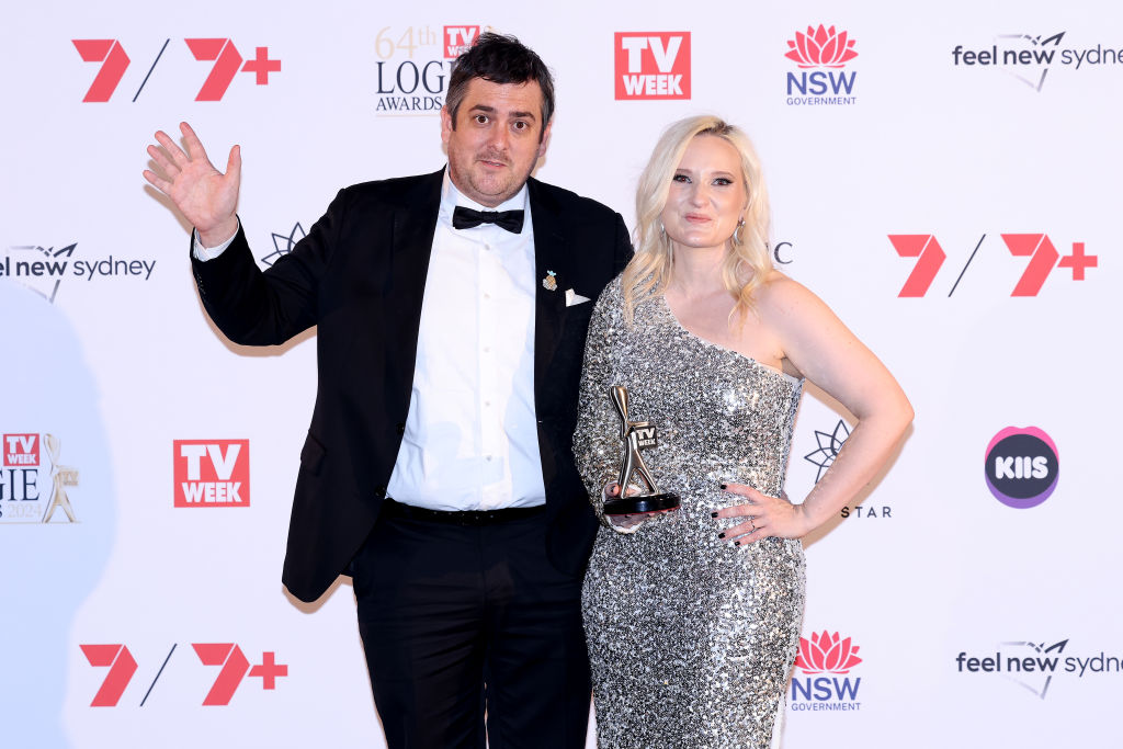 Man in tuxedo and woman in silver dress holding award at TV WEEK event backdrop.