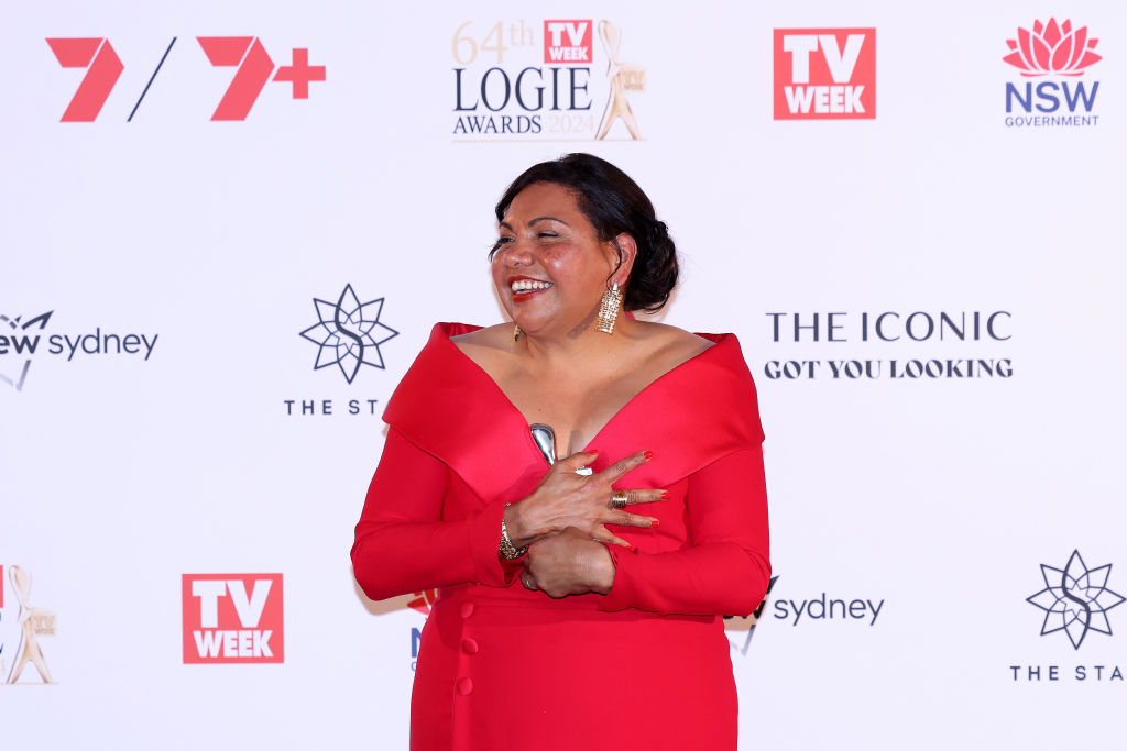 A woman in a red dress smiles on the Logie Awards red carpet, logos in background.