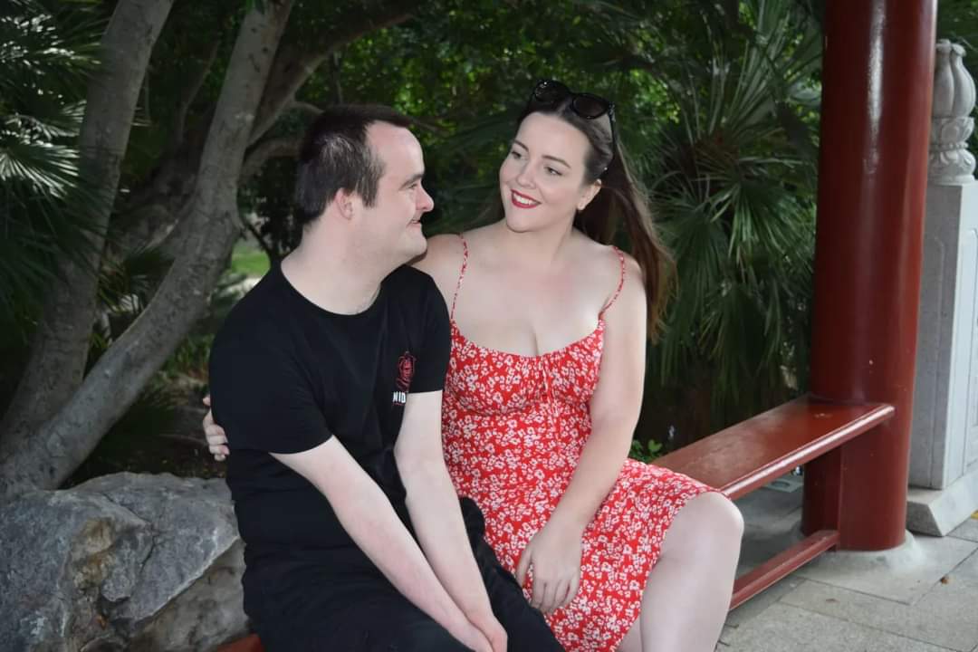 Tom and his sister and carer, Amanda, sit on a garden bench. Tom wears black jeans and t-shirt. Amanda wears a red floral print dress with shoestring straps.