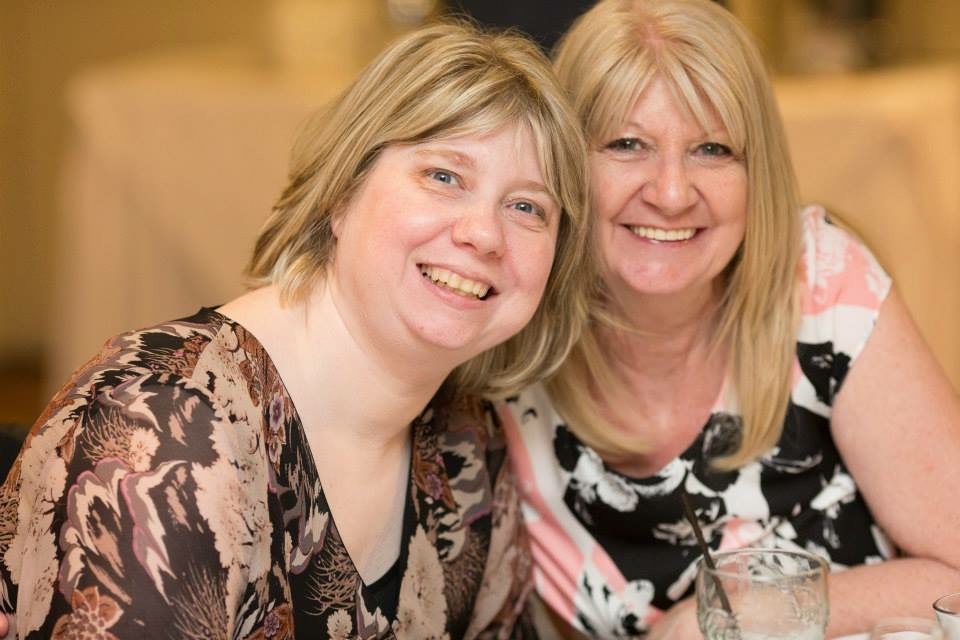 Lee-Ann and Dee smile for a photograph. Both have blonde hair and wear print dresses in shades of black, brown and pink.
