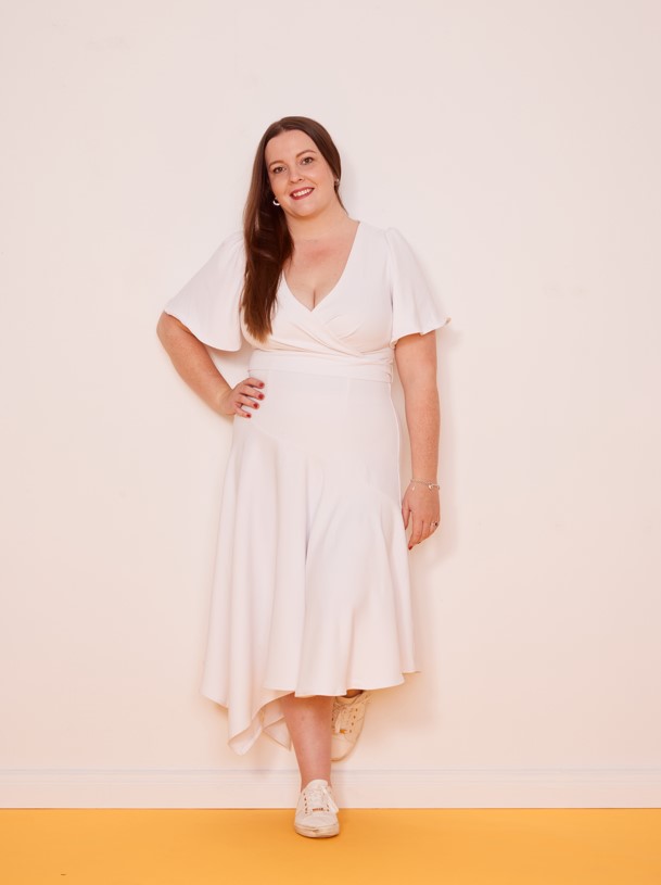 Amanda stands in a stodio with a yellow floor and pale pink wall. She wears a white, calf-length wrap dress. Her hair is long and mid-brown.