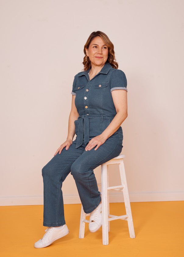 Tairyn sits on a white stool in the photographic studio. She wears a blue denim jumpsuit.