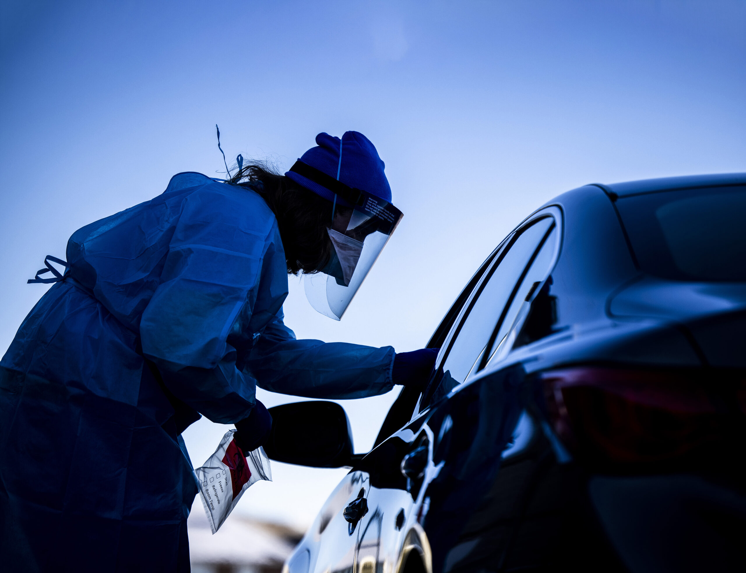 A health worker in full PPE stands beside a car holding a plastic bag and leaning towards the window. It is dusk.