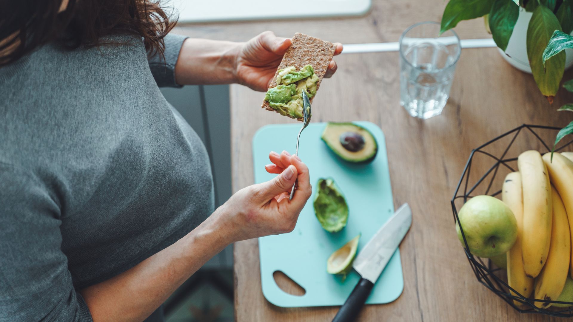 Top view of unrecognisable pregnant woman wearing grey making healthy toast with avocado in her kitchen.